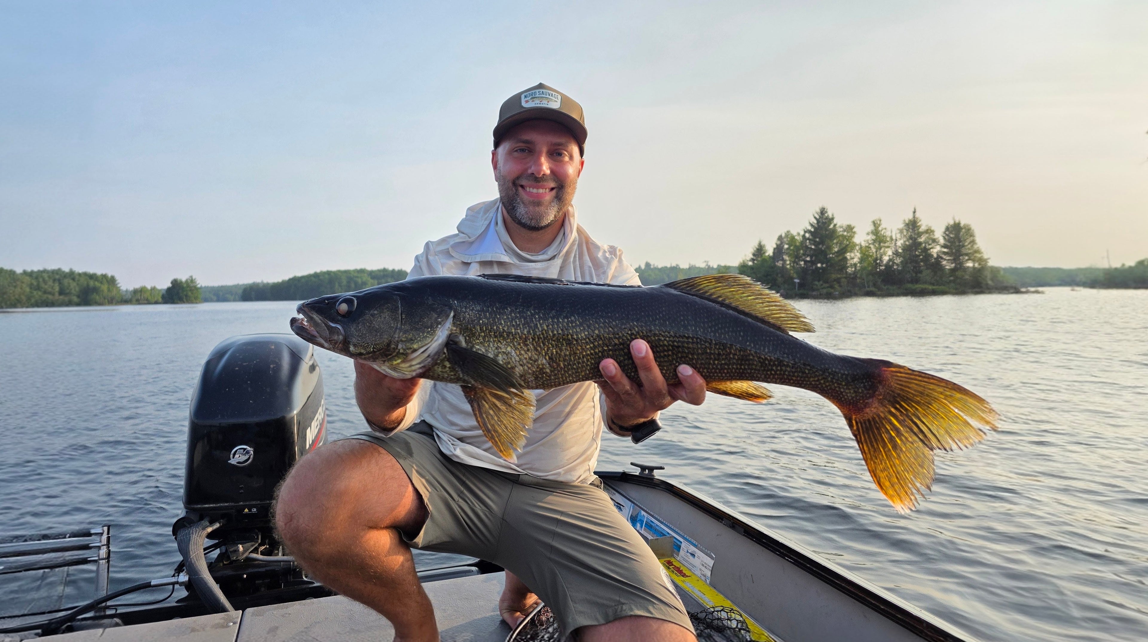 Man holding a large fish on a boat with a lake and trees in the background