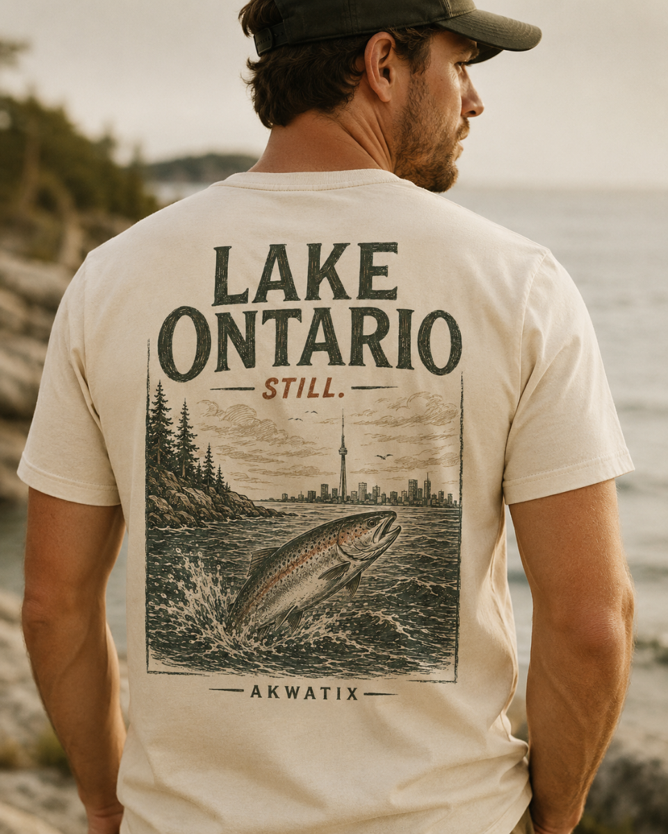A man wearing a beige t-shirt with a large back graphic that reads 'LAKE ONTARIO STILL. AKWATIX' above and below an illustration of a leaping fish and a shoreline cityscape.