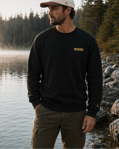 A man wearing a black crewneck sweatshirt with a small gold 'HARVEY' logo on the chest, paired with a beige baseball cap and olive cargo pants, standing on a rocky lakeshore at sunrise.