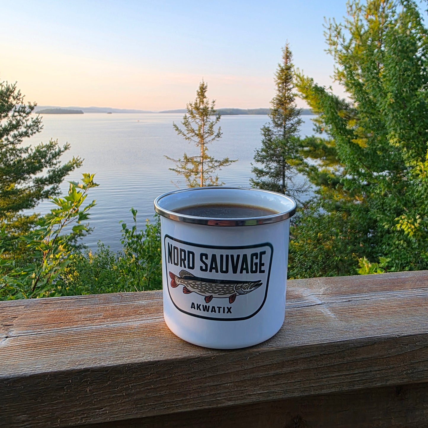 A white enamel camping mug with a fish logo and the words 'WORD SAUVAGE' and 'AKWATIX' sits on a wooden railing with a scenic lake and forest background.